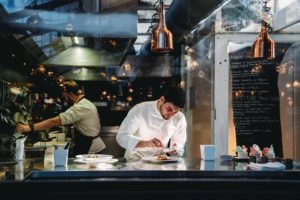 View through clean window of chef preparing beautiful plate of food in professional kitchen